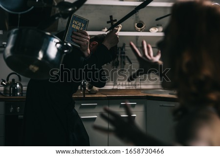 selective focus of evil demon with levitating cookware and exorcist with cross and bible in kitchen Royalty-Free Stock Photo #1658730466