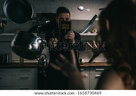 selective focus of demon with levitating cookware and exorcist with cross and bible in kitchen Royalty-Free Stock Photo #1658730469