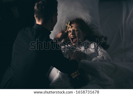 male exorcist with bible and cross standing over demoniacal screaming girl in bed Royalty-Free Stock Photo #1658731678