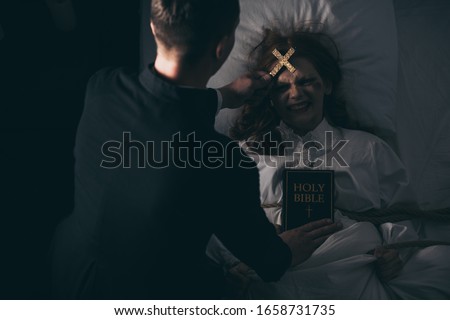 exorcist with bible and cross standing over demonic obsessed girl in bed Royalty-Free Stock Photo #1658731735