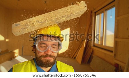 CLOSE UP, PORTRAIT: Wooden board falls on a young worker's head. Small plank falls from above on an unsuspecting contractor's head. Protective hard hat saves Caucasian builder's from a concussion.