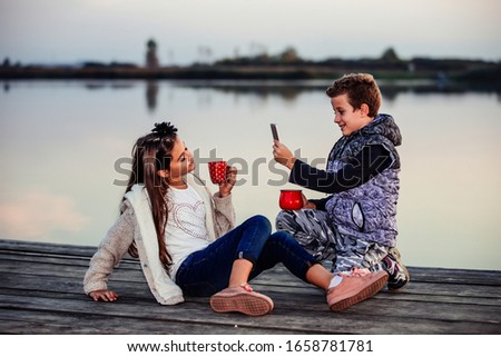 Two young cute little friends, boy and girl  having fun and drinking tea while sitting by the lake in the evening. Boy taking a photo with phone of his friend. Friendship