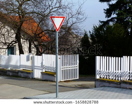 White metal picket fence, gate and sidewalk. house detail with white stucco elevation and brown clay roof in winter season. triangular white and red yield road traffic sign. pale blue sky. bare trees,