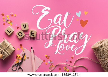 top view of valentines decoration, scissors, gift box, twine and love lettering on wooden cubes on pink background with eat, pray, love lettering