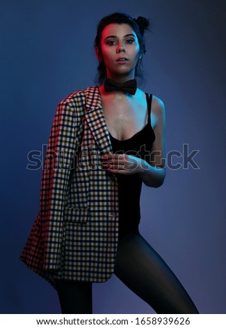 Cabaret, dancer and holidays concept - Cute young girl in the bow tie, black body bikini hold jacket on the shoulder. ?olorful neon light on dark background