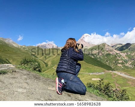 young female takes pictures with her camera of kazbek mountain and white clouds over it, summer in caucasian mountains, 