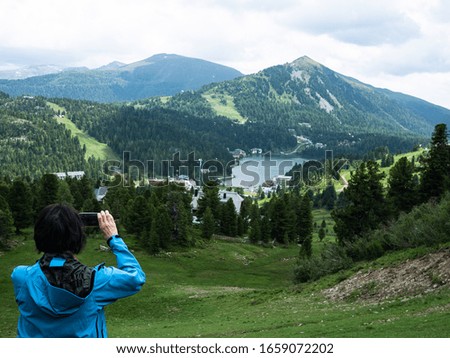 woman in blue jacket is taking a picture with her smartphone of a green alpine landscape with a lake