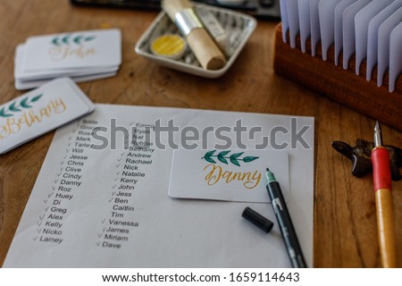 Wedding calligraphy - guest list and handwritten place cards with names. Golden writing with green flourish on white paper. Gold paint, nib pen, card holder. Royalty-Free Stock Photo #1659114643
