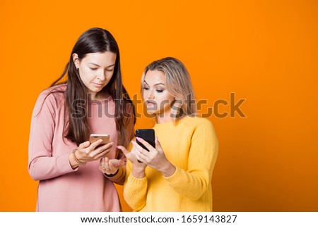 Two cheerful friends communicate and show each other something in phones, live communication, modern technologies. Two friendly young women on a yellow background