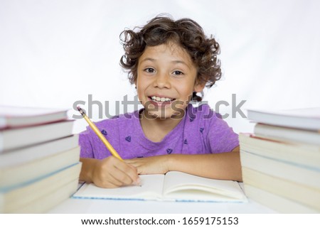 Happy boy between 8 and 10 years old sitting at a desk studying while holding a pencil with textbooks on his table and with a white background