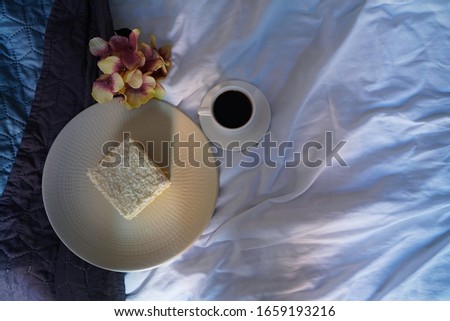 Breakfast in the bed. On the wite sheets the cup of coffe, flowers and the plate with cake