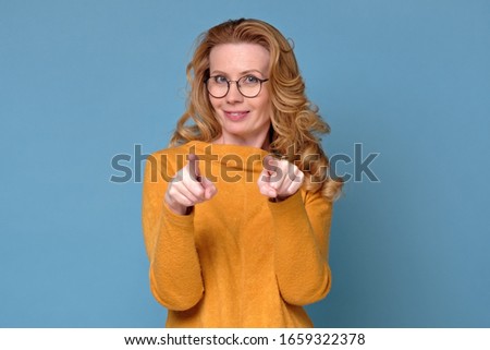 Happy mature woman in glasses pointing at you. I choose you. Studio shot on blue wall.