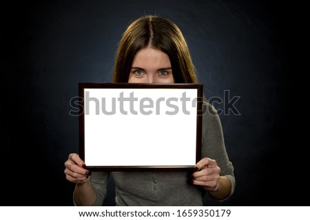 Portrait of a young woman 25-30 years old holding a white text frame on a dark background, shyly hiding her face, smiling