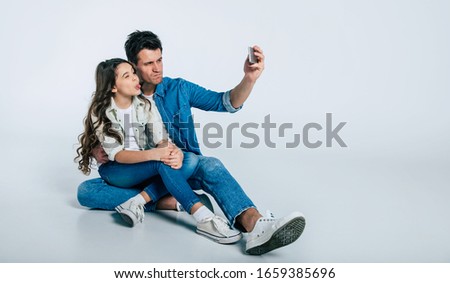 A new selfie picture. Full-length photo of a happy father in a denim outfit, who is sitting on the floor with his cute little child and grimacing, while making selfies with her.