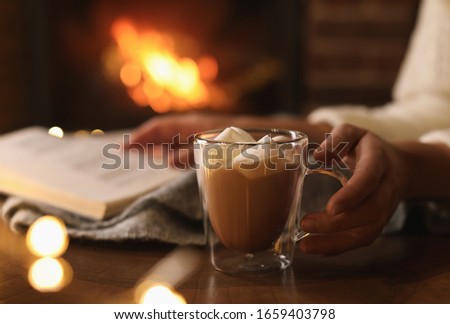 Woman with cocoa and book near burning fireplace indoors, closeup