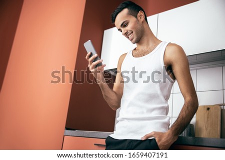 Happy young man in t-shirt looking at mobile phone screen while resting at home stock photo