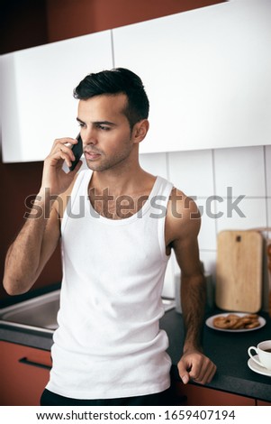 Waist up of young guy in white t-shirt using mobile phone while looking away at the kitchen stock photo