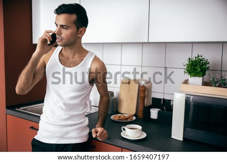 Waist up of man in t-shirt holding mobile phone while looking away at the kitchen stock photo