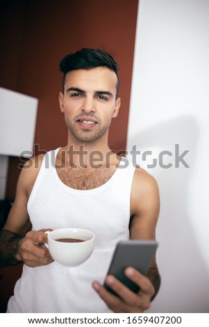Happy guy in white t-shirt using smartphone while holding coffee at home in the morning stock photo