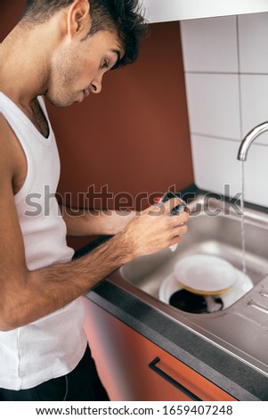 Young guy using sponge while washing the cup under running water at home stock photo