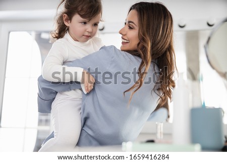 Waist up of smiling pretty mom playing with her small daughter in room stock photo