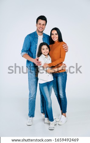 Happy together. Full-length family picture of mom, dad and their cute daughter in casual clothes, standing in front, smiling and hugging together.