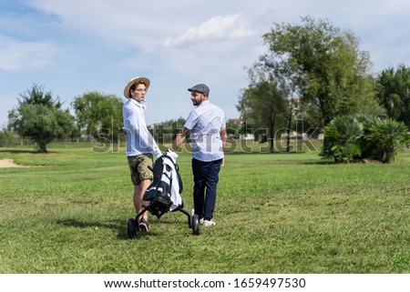 Couple of men in hats walking backwards through a golf course with a cart to carry the equipment while turning their heads behind