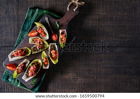 Endive salad boats filled with pico de gallo over minced beef served on a rustic cutting board on a dark wooden background, horizontal orientation, top view, copy space