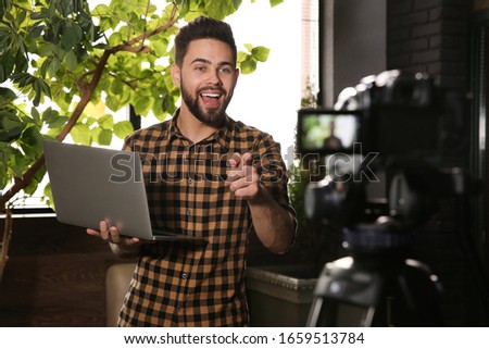 Young blogger with laptop recording video on camera indoors