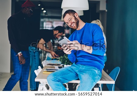 Cheerful caucasian hipster guy in eyewear for vision correction laughing at funny photos on digital tablet during work break, happy millennial male 20s having fun watching videos from social networks