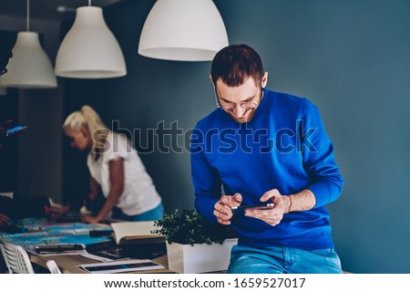 Cheerful young 20s male in eyewear satisfied with new app on digital tablet playing online games on free time indoors, happy caucasian hipster man watching videos on touchpad from social networks