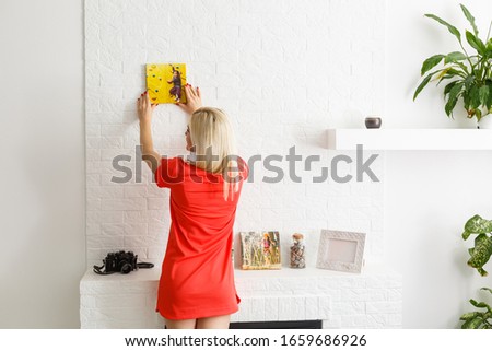 A happy young blonde woman is holding a large wall canvas at home