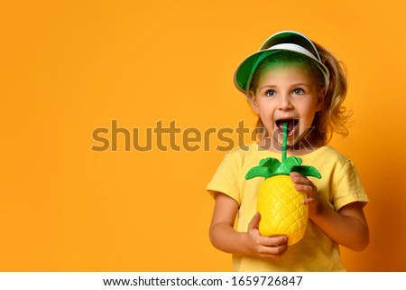 Little smiling cute blond girl in yellow t-shirt and hat drinking fresh healthy fruit juice from straw over yellow background. Healthy lifestyle and clean eating concept