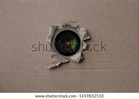 eye of the camcorder looks through a torn hole in an empty brown cardboard, craft paper, concept of secrecy, covert video surveillance, tracking, industrial espionage, blank for designer, copy space