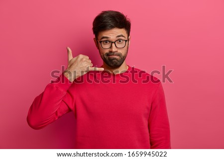 Serious looking unshaven European man makes call me back gesture, keeps always in touch, wears transparent spectacles and red sweater, asks for telephone number, isolated on pink background.
