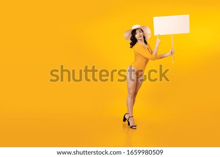 Young brunette woman with pin-up hairstyle in a yellow bathing suit and white hat holds a white poster in her hands and poses on a yellow background