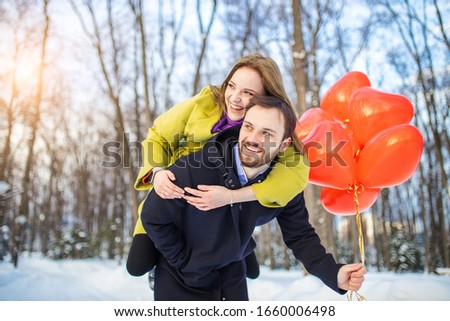 couple having promenade. romantic man and woman in coats dating, hold red air balloons in hands, they look happy together. attractive lady jumped on man's back and laugh. outdoors. relationships