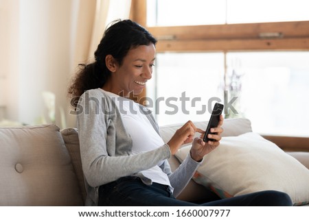 Smiling young African American woman sit on couch at home pose make self-portrait picture on modern cellphone, happy millennial biracial female relax on sofa take selfie on cellphone camera