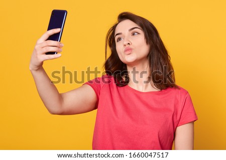 Indoor shot of happy flirting young girl taking pictures of herself through cell phone, charming female over yellow background, darkhaired girl wearing red casual t shirt. Modern technology concept.