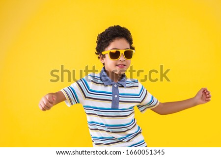 Dark skinned curly smiling boy in blue striped t-shirt is listening music and dancing on bright yellow background. Fashionable black child in glasses is having fun. Modern children concept.