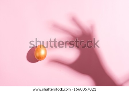Creative background from shadows of woman's hand with Easter golden egg against a pastel pink background.