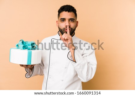 Young latin baker man holding a cake isolated keeping a secret or asking for silence.