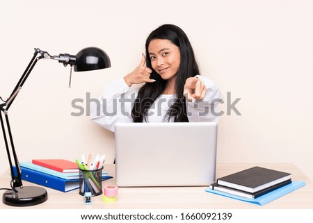 Student asian girl in a workplace with a laptop isolated on beige background making phone gesture and pointing front