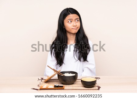Teenager Asian girl eating asian food isolated on beige background making doubts gesture looking side