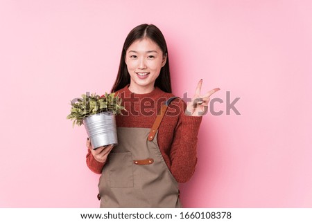Young chinese gardener woman holding a plant isolated joyful and carefree showing a peace symbol with fingers.
