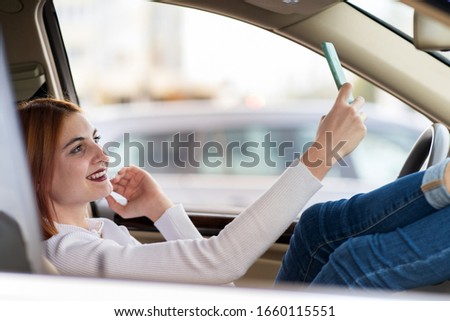 Young redhead woman driver taking selfies with her mobile phone sitting behind the wheel of the car in rush hour traffic jam.