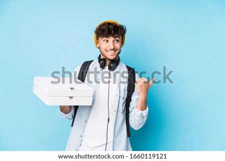 Young arab student man holding pizzas isolated points with thumb finger away, laughing and carefree.