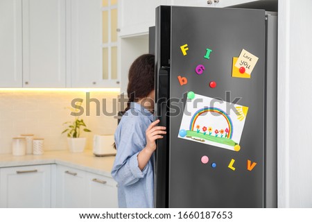 Woman opening refrigerator door with child's drawing, notes and magnets in kitchen Royalty-Free Stock Photo #1660187653