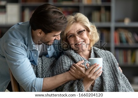 Happy middle-aged mother relax in chair drink tea enjoy family weekend reunion with grown-up son, smiling senior 70s mom rest at home spend time with caring adult man child, bonding concept Royalty-Free Stock Photo #1660204120