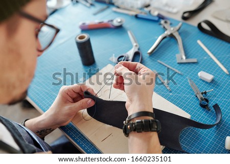 Contemporary craftsman sewing two parts of black leather together while holding them over wooden board on table